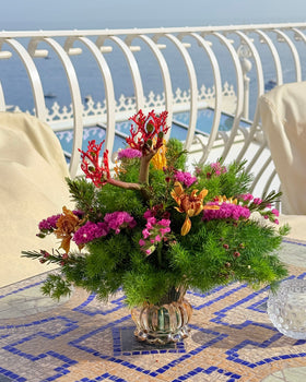 Centrotavola con fiori fucsia, arancioni e ramificazioni rosse in vaso di vetro su tavolo a mosaico, con il mare sullo sfondo.